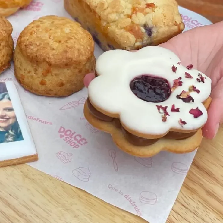 Alfajor con forma de flor, glaseado en blanco, con mermelada en el centro y pétalos de rosa, sostenido en la mano sobre una tabla con otras masas dulces